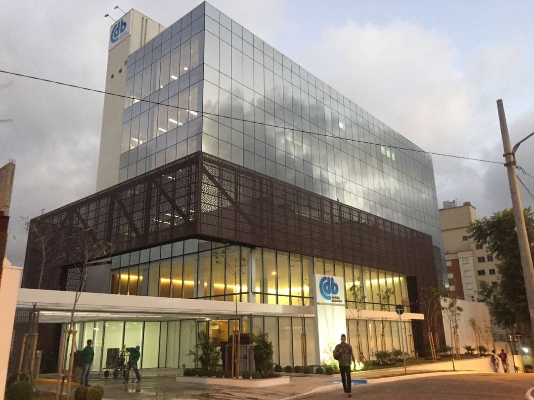 Fachada envidraçada de unidade do CDB ao entardecer, com brise metálico e térreo iluminado.