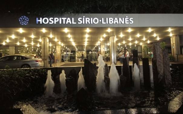 Fachada iluminada da entrada principal do Hospital Sírio-Libanês, em São Paulo, com espelho d'água em primeiro plano.